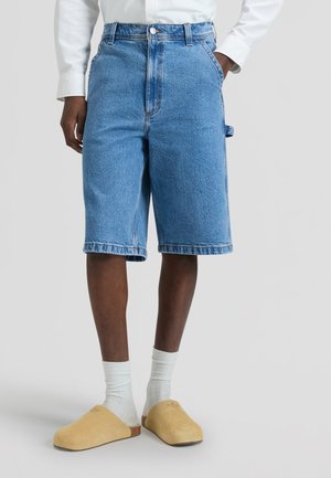 Isik, kes kannab siniseid denimist põlvpükse, valgeid sokke, beeže libisemisjalanõusid ja valget pikavarrukalist särki, seisab.