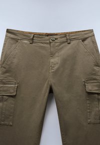 Khaki cargo bukser med flere sidelommer, en knaplukning og en glat tekstur. Har et kontrastfarvet orange detalje på taljen.
