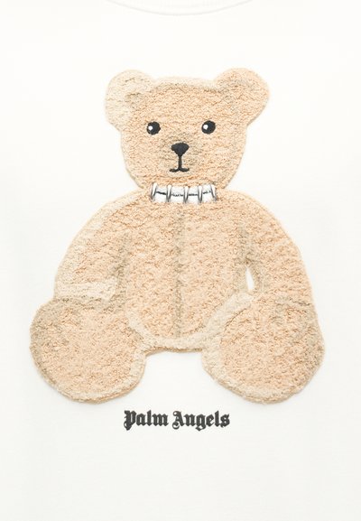 Μπεζ αρκουδάκι teddy με υφή γούνας και ασημένιο κολάρο με ακίδες πάνω από το κείμενο "Palm Angels" σε λευκό φόντο.