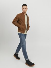 Chaqueta bomber de ante marrón, suéter ligero, jeans azules y botas de tobillo negras con suelas marrones. Modelo caminando contra un fondo simple.