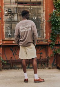 Un homme portant un short beige, une chemise à manches longues marron avec du texte au dos, des chaussettes blanches et des chaussures marron, se tient face à une fenêtre grillagée couverte de graffitis et de vignes.