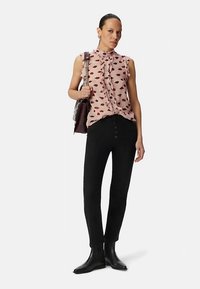 Kwiecista bluzka z nadrukiem czerwonych ust na jasnoróżowym tle, z falbankowanym kołnierzykiem, w połączeniu z czarnymi dopasowanymi spodniami i czarnymi botkami, trzymająca torebkę.