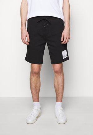 Zwarte katoenen shorts met een trekkoord in de taille, zijzakken en een wit "POLO" logo op het linkerbeen. Gecombineerd met witte sneakers en sokken.