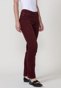 Vrouw in slim-fit bordeauxrode corduroybroek, witte mouwloze top en zwarte platte schoenen met gespsluiting, staande op een effen achtergrond.
