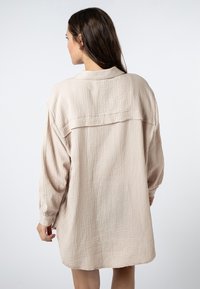 Lichtbeige oversized hemdjurk gemaakt van geborsteld stof, met een kraag, lange mouwen en een losse pasvorm die halverwege de bovenbenen valt.