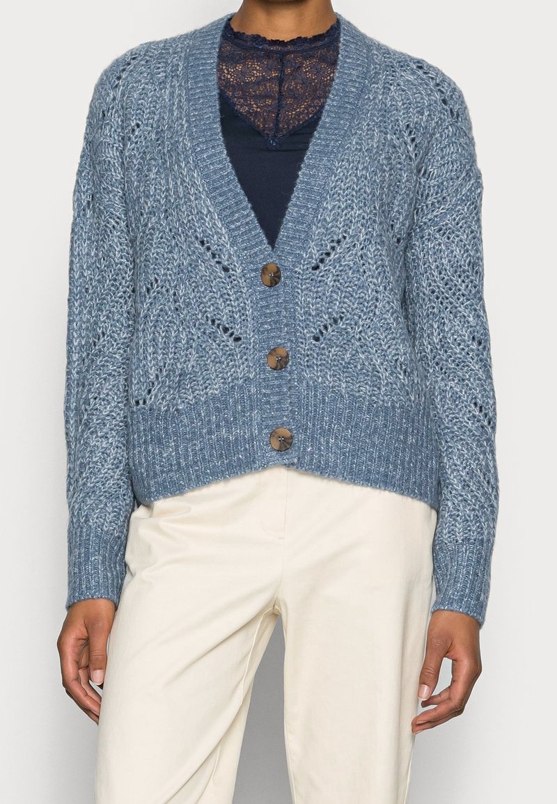 Personne portant un cardigan bleu texturé boutonné par-dessus un haut en dentelle marine et un pantalon crème, debout avec les bras détendus le long du corps.