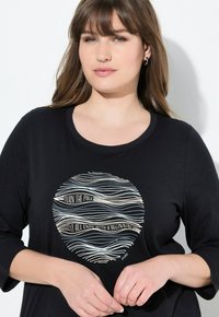 Un haut noir à manches longues arborant un graphique circulaire avec des lignes ondulées et le texte "TOURNER LA PAGE / TOUT SE TERMINE PAR UN DÉBUT" en crème et bleu.
