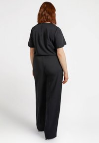 Mono negro con mangas cortas, cintura elástica y diseño de pierna ancha. Tejido suave con un detalle de abertura en la parte posterior del cuello.