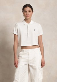 Witte cropped polo shirt met een klassieke kraag, voorzien van een knoopsluiting en een klein donker logo. Gekoeld met witte cargobroeken.