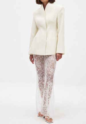 Balta struktūrizuota švarkas su aštriu apykakle, derinama su ilgomis skaidriomis nėrinių kelnėmis, kuriose yra gėlių raštai ir prigludusi siluetas.