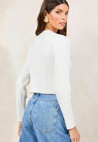 Witte kabelgebreide cropped trui met lange mouwen, gecombineerd met lichtblauwe high-waisted jeans. Vloeiende stof met een gestructureerd ontwerpdetail.