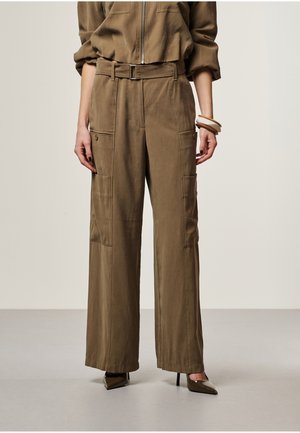 Vrouw draagt bruine wijd uitlopende cargobroek met riem, gecombineerd met donkere schoenen met puntige neus en armbanden om haar pols.