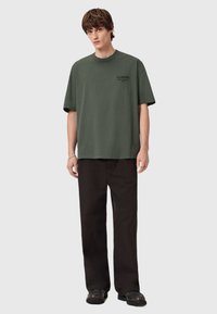 Yksi koko oliivinvihreä t-paita puuvillasta, jossa on pyöreä pääntie ja lyhyet hihat, yhdistettynä mustiin löysiin housuihin.