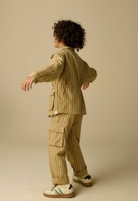 Enfant aux cheveux bouclés portant une veste rayée beige et un pantalon cargo, debout avec les bras légèrement levés devant un fond uni.