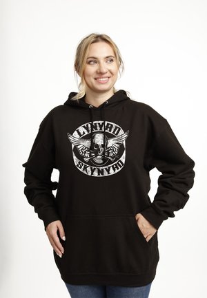 Uśmiechnięta kobieta w czarnej bluzie Lynyrd Skynyrd z logo czaszki i skrzydeł, ręce w kieszeniach, jasne włosy związane do tyłu, stoi na białym tle.
