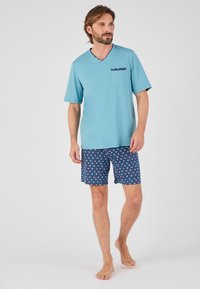 Lichtblauw T-shirt met korte mouwen en V-hals, voorzien van een zak. Marineblauwe shorts met een klein ankerpatroon in het wit en rood. Model is blootsvoets.