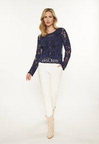 Suéter de encaje azul marino con patrones intrincados y cierre de botones, combinado con pantalones ajustados blancos y botas de tobillo beige.