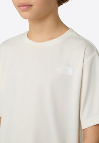 Oseba, oblečena v belo majico kratkih rokavov znamke North Face, z vidnim izrezom na vratu in logotipom na levi strani prsnega koša.