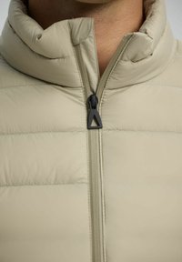 Beige pufferjack met hoge kraag, voorzien van horizontale gestikte banen en een centrale rits met een driehoekige trekker. Soepele, zachte textuur.