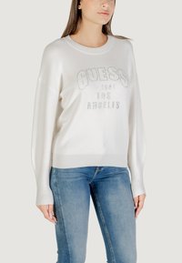 Witte gebreide trui met geribbelde boorden en onderrand, voorzien van het 'GUESS'-logo in een studded ontwerp. Gecombineerd met lichtblauwe skinny jeans.