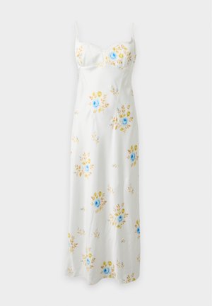 Mujer con cabello largo y negro, vistiendo un vestido midi blanco con estampado floral azul y amarillo y tacones puntiagudos en color crema, de pie frente a un fondo liso.