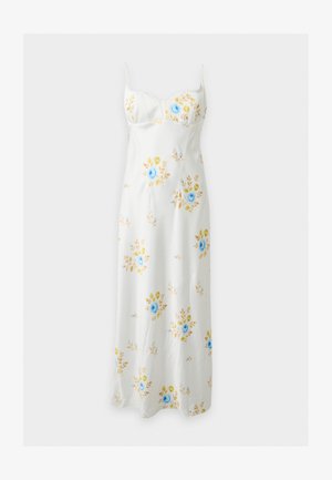 Mujer con cabello largo y negro, vistiendo un vestido midi blanco con estampado floral azul y amarillo y tacones puntiagudos en color crema, de pie frente a un fondo liso.