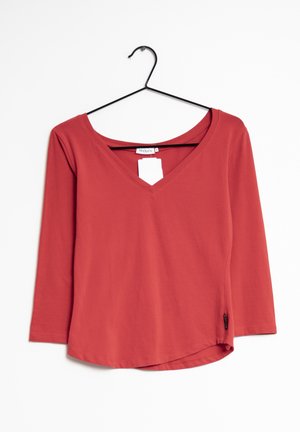 Maglia rossa a maniche lunghe con scollo a V, realizzata in tessuto morbido, con orlo curvo e una piccola etichetta del marchio sul lato. Appesa a un gancio nero.