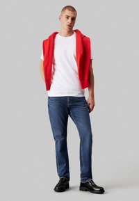 Sudadera sin mangas roja sobre una camiseta blanca, combinada con jeans azules y zapatos negros. Diseño simple sin patrones o acentos visibles.