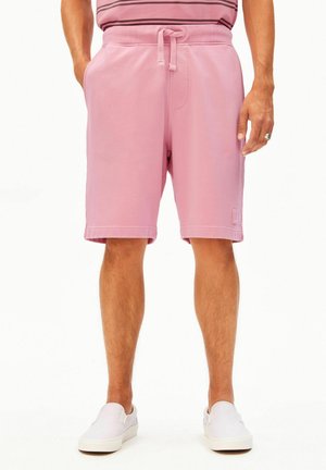 Mann trägt pastellrosa Shorts mit Kordelzug und weiße Slip-On-Schuhe, steht mit entspannten Händen an den Seiten.