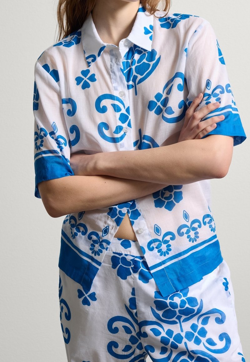 Witte shirt met blauwe bloemmotieven, korte mouwen en een button-down kraag. Kenmerkt zich door een uitsparing onder de knopen en een contrasterende zoom.