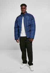 Navy blue puffer jacket with quilted design, high collar, and snap buttons, worn over a white T-shirt and black cargo pants. White sneakers.