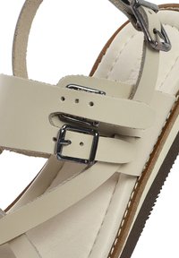 Beige leren sandaal met verstelbare banden en zilveren gespen, een gevoerde binnenkant en een bruine rubberen zool met witte stiksels.