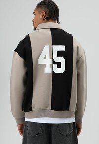 Oversized jack met kraag in beige en zwart kleurvlakken. Voorzien van een grote witte "45" op de achterkant en een geribde zoom. Zachte stoftextuur.