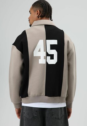 Oversized jack met kraag in beige en zwart kleurvlakken. Voorzien van een grote witte "45" op de achterkant en een geribde zoom. Zachte stoftextuur.
