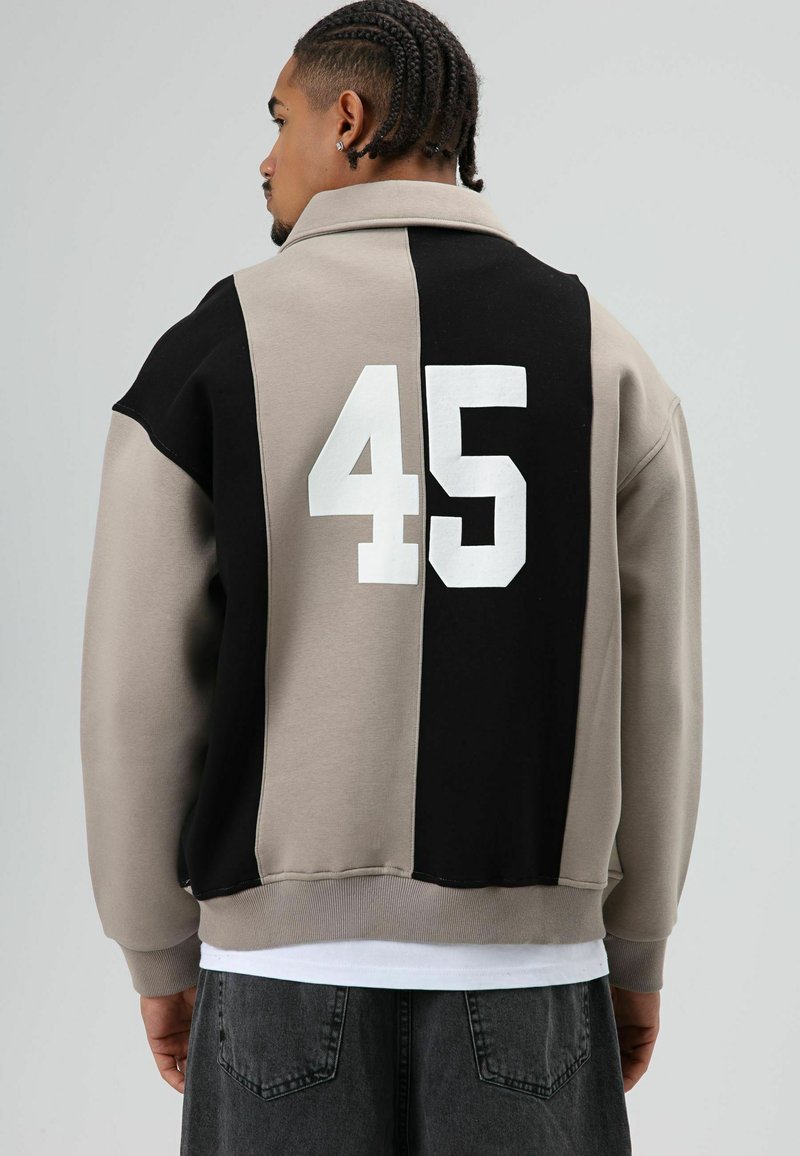 Oversized jack met kraag in beige en zwart kleurvlakken. Voorzien van een grote witte "45" op de achterkant en een geribde zoom. Zachte stoftextuur.