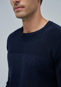 Nahaufnahme eines Mannes, der einen strukturierten, navyblauen Strickpullover mit einem dezent gestickten Logo auf der Brust trägt.