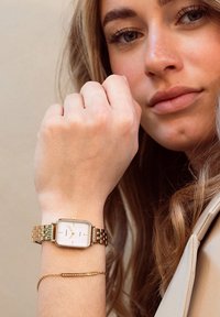 Montre rectangulaire en or avec un bracelet en maillon métallique, dotée d'un cadran blanc et d'accents dorés. Un bracelet fin en or est porté en parallèle au poignet.