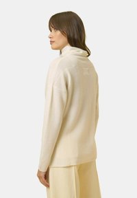 Cremfarbener Rollkragenpullover mit lockerer Passform, aus texturiertem Strickstoff und mit einem kleinen Etikett auf der Rückseite. Kombiniert mit hellen Hosen.