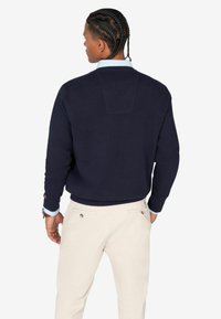 Felpa navy con fondo e polsini a costine. La parte posteriore presenta un design a pannello contrastante. Abbinata a pantaloni di colore chiaro.