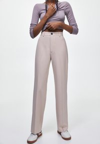 Pantalon beige taille haute en tissu lisse. Doté d'une fermeture à bouton, de poches latérales et d'une coupe droite. Porté avec des baskets blanches.