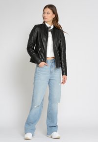 Schwarze Lederbikerjacke mit gesteppten Schultereinsätzen, kombiniert mit hellblauen, zerrissenen Jeans und weißen Sneakers. Schlichter Hintergrund.
