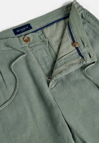 Pantalon en coton vert avec fermeture éclair partiellement ouverte, bouton, taille à cordon de serrage et étiquette de ceinture intérieure visible "MR MARVIS Amsterdam".