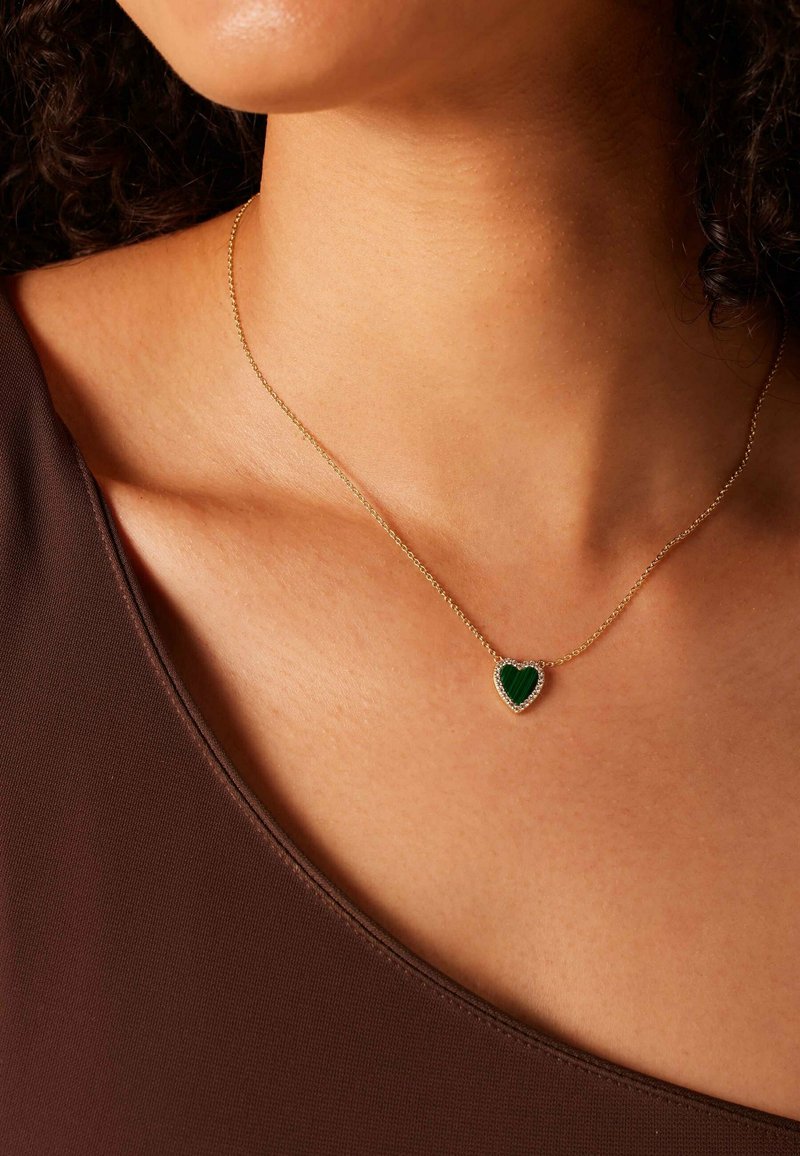 Gouden ketting met een hartvormige hanger met een groene steen, omringd door kleine heldere stenen, die op een bruine kledingstuk ligt.