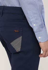 Pantalones azul marino con un panel de tela gris en el bolsillo trasero, con un diseño de perro bordado en marrón. Camisa de algodón texturizada con un patrón de puntos.