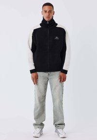 Un jeune homme se tient face à l'avant, portant une veste zippée noire et blanche, un jean gris clair et des baskets blanches sur un fond uni.
