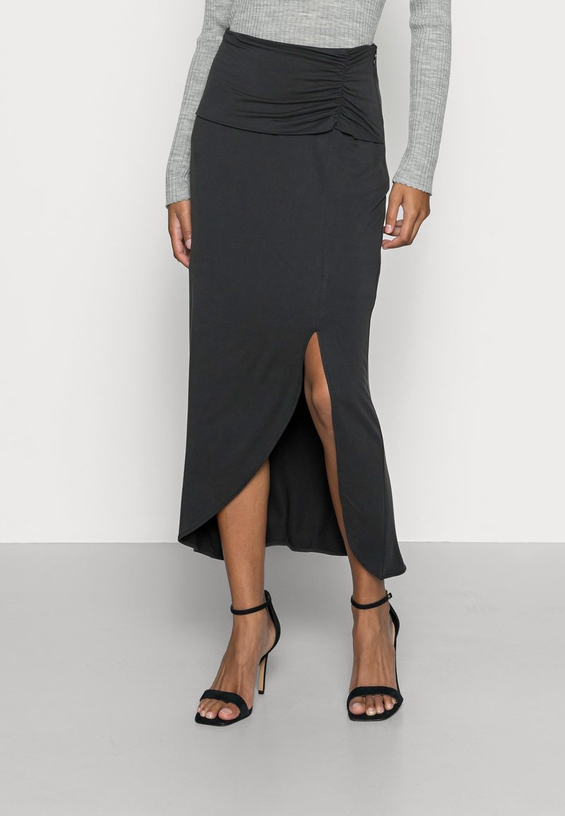 Schwarze, figurbetonte Midirock mit drapiertem Bund, seitlichem Schlitz und asymmetrischem Saum. Getragen mit schwarzen, Riemchen-High-Heels.