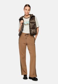 Leopard print padded vest, light-colored graphic t-shirt, brown trousers with side stripes, and black chunky sole shoes.