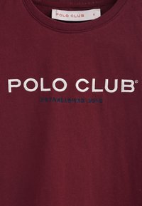 Camiseta de algodón burdeos con cuello redondo. Presenta "POLO CLUB" bordado en blanco y "ESTABLISHED 2012" en azul. Etiqueta de talla visible.