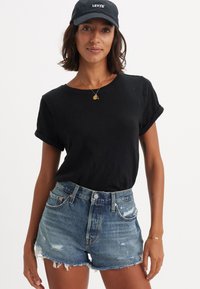 Camiseta negra de manga enrollada combinada con pantalones cortos de denim azul desgastado con dobladillos deshilachados y detalles envejecidos. Gorra negra con el logotipo de "Levi's".