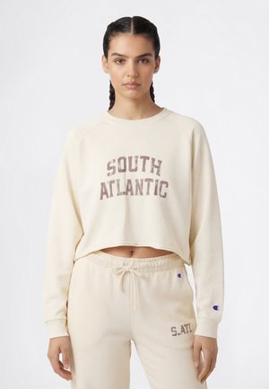 Donna con capelli intrecciati che indossa una felpa color crema con la scritta "South Atlantic" e pantaloni della tuta abbinati con coulisse e piccole toppe con logo.